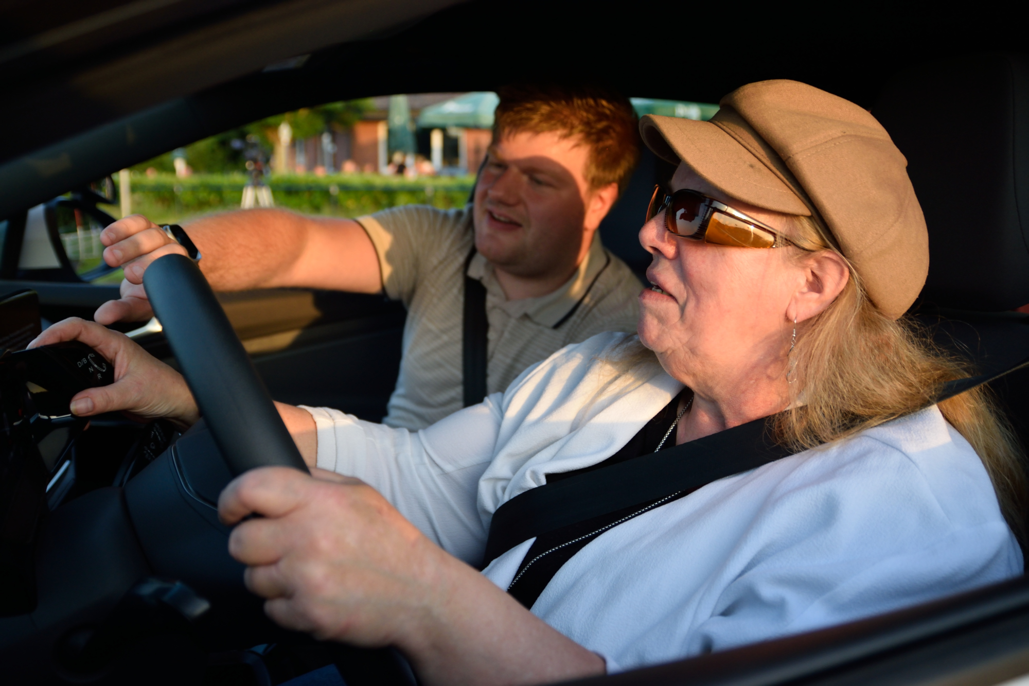 Eine ältere Frau mit Sonnenbrille und Hut hsitzt auf dem Fahrersitz eines Autos und hält das Lenkrad fest, während ein Fahrlehrer auf dem Beifahrersitz auf die Bedienelemente am Armaturenbrett zeigt. Eine ältere Frau mit Sonnenbrille und Hut hsitzt auf dem Fahrersitz eines Autos und hält das Lenkrad fest, während ein Fahrlehrer auf dem Beifahrersitz auf die Bedienelemente am Armaturenbrett zeigt.