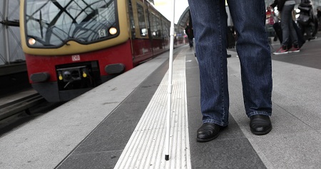 Das Foto zeigt eine Person mit einem Blindenstock an einem Bahnhgleis. Die Person orientiert sich über ein im Boden integrieretes Leitsystem während ein Zug einfährt