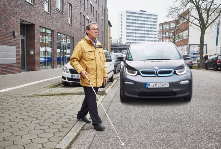 Ein Mann mit Langstock will die Straße vor einem E-Auto überqueren