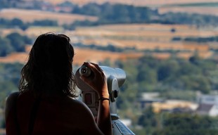 Eine Frau steht vor einem Fernglas, im Hintergrund eine verschwommene Landschaft mit Bäumen und Feldern