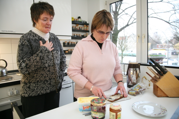 Eine Frau schmiert Butter auf eine Scheibe Brot, beobachtet von der Reha-Lehrerin